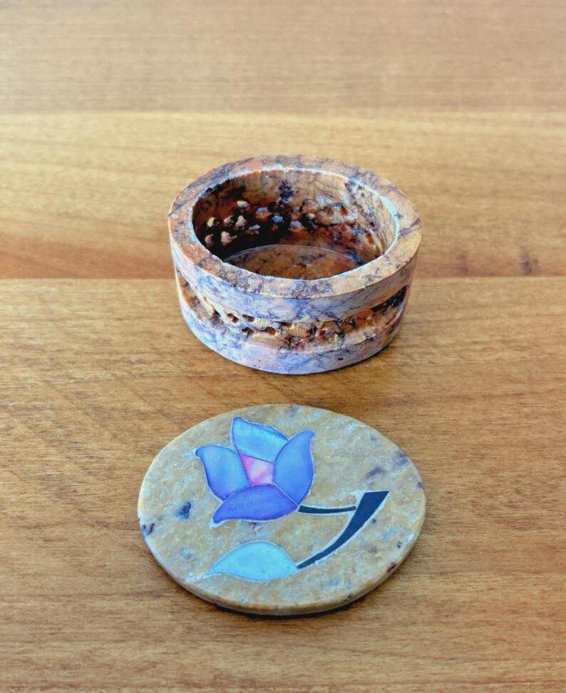 May include: A small, round, beige stone box with a matching lid. The lid features a blue flower design with a black stem. The box has a carved design around the sides. The box and lid are on a wooden surface.