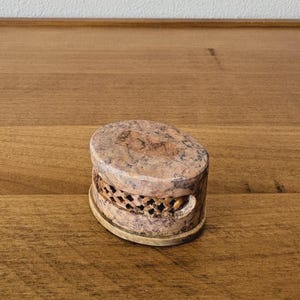 May include: A small, oval-shaped stone box with a lid. The box is tan and brown with a speckled pattern. The sides of the box have a carved design. The box is sitting on a wooden surface.