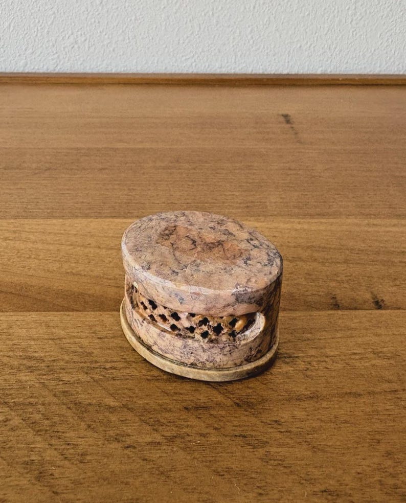 May include: A small, oval-shaped stone box with a lid. The box is tan and brown with a speckled pattern. The sides of the box have a carved design. The box is sitting on a wooden surface.