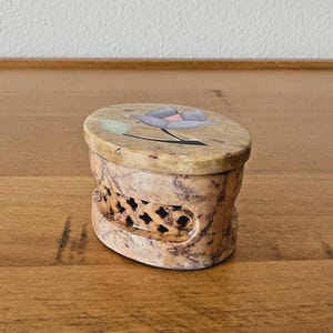 May include: Oval-shaped, beige stone box with a carved floral design on the lid. The lid features a purple flower with green and black accents. The sides of the box have a carved, openwork pattern.
