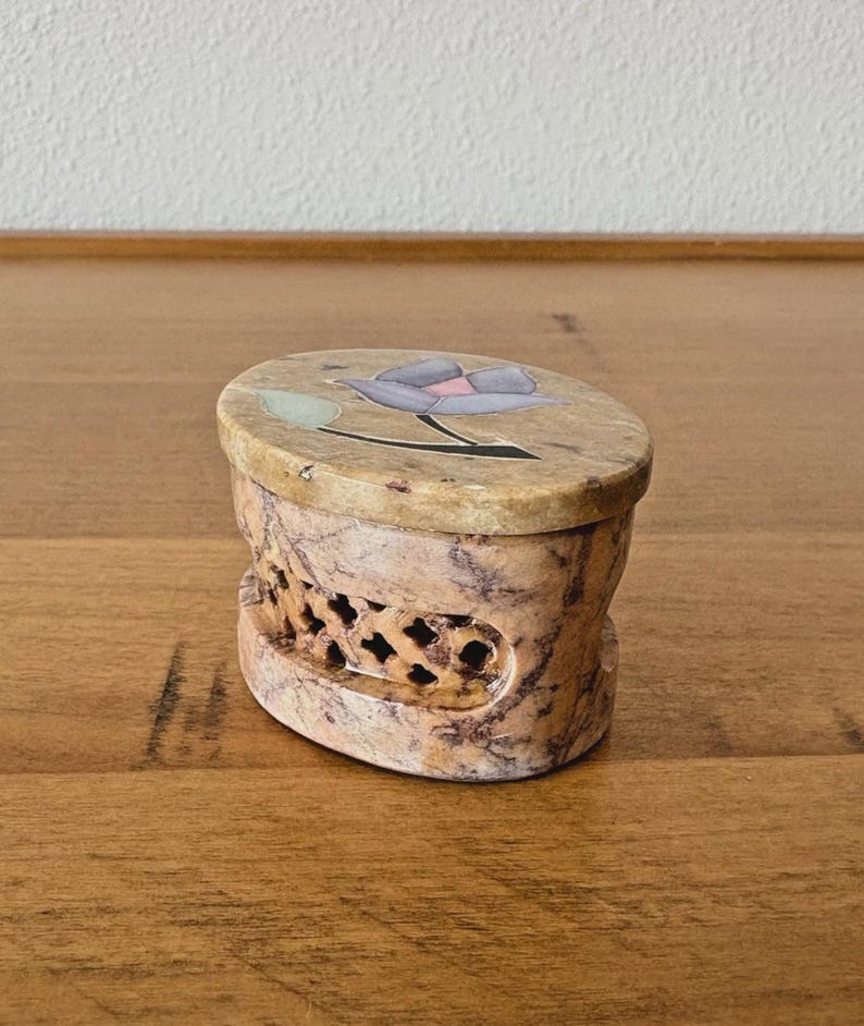 May include: Oval-shaped, beige stone box with a carved floral design on the lid. The lid features a purple flower with green and black accents. The sides of the box have a carved, openwork pattern.
