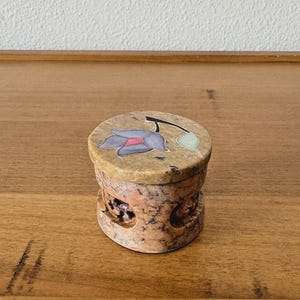 May include: A small, round, stone box with a lid. The lid features a floral design with purple and pink petals. The box has a carved, textured body with a natural, earthy color palette.