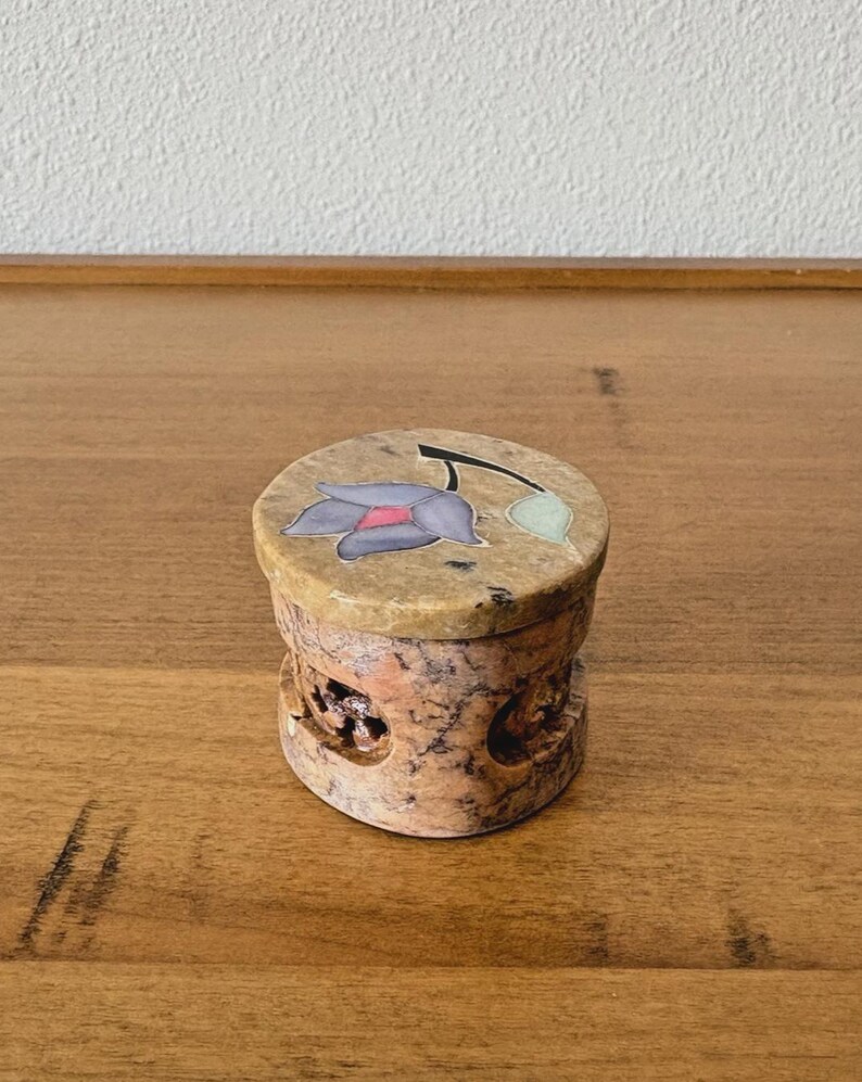 May include: A small, round, stone box with a lid. The lid features a floral design with purple and pink petals. The box has a carved, textured body with a natural, earthy color palette.