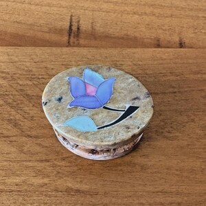 May include: A round, beige stone box with a blue and purple floral design on the lid. The flower has a black stem and a light blue leaf. The box is sitting on a wooden surface.