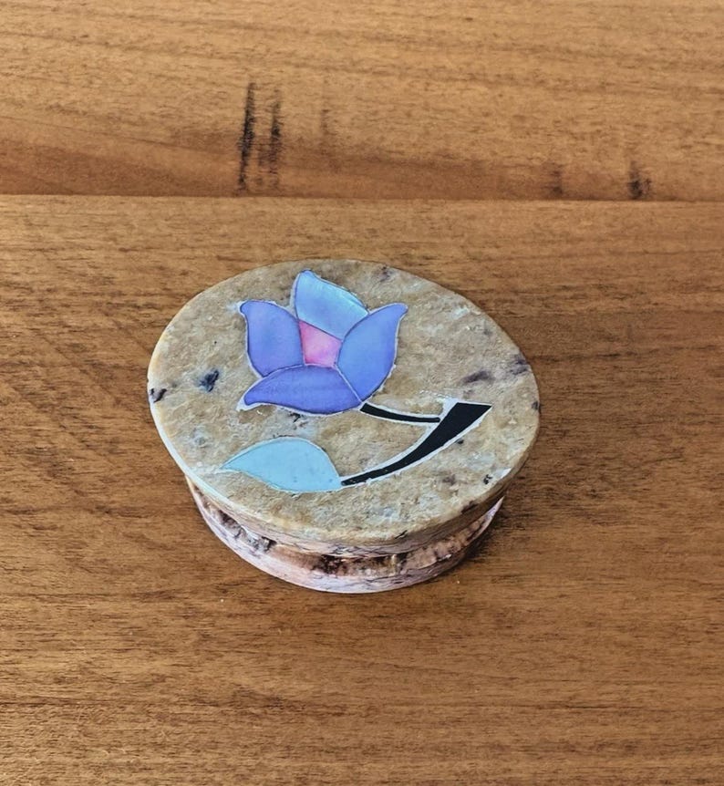 May include: A round, beige stone box with a blue and purple floral design on the lid. The flower has a black stem and a light blue leaf. The box is sitting on a wooden surface.