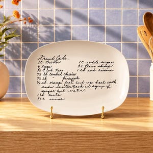 May include: An oval white ceramic platter featuring black cursive text that lists a fruit cake recipe including ingredients like butter, eggs, candied cherries, pineapple, orange peel, nuts, and wine, displayed on a small gold metal stand.