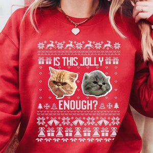 May include: Red sweatshirt with a festive design. The design includes two cat faces, text that reads "IS THIS JOLLY ENOUGH?", and holiday-themed patterns. The sweatshirt is a crew neck style.