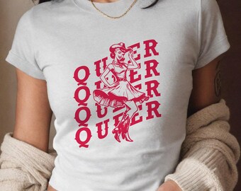 May include: A white t-shirt with a red graphic of a woman in a cowboy hat and dress. The text "QUEER" is repeated five times in a vertical stack.