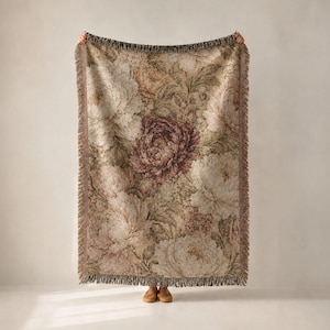May include: A rectangular woven throw blanket featuring a detailed floral pattern with large, layered peony-like blossoms in shades of muted pink, deep burgundy, cream, and sage green, finished with a decorative fringed edge on all sides.