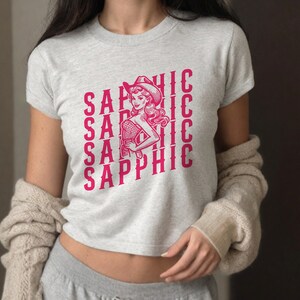 Może przedstawiać: Szara, kr&oacute;tka koszulka z r&oacute;żowym nadrukiem przedstawiającym kobietę w kowbojskim kapeluszu trzymającą nap&oacute;j. Tekst "SAPPHIC" jest powt&oacute;rzony na koszulce wiele razy.