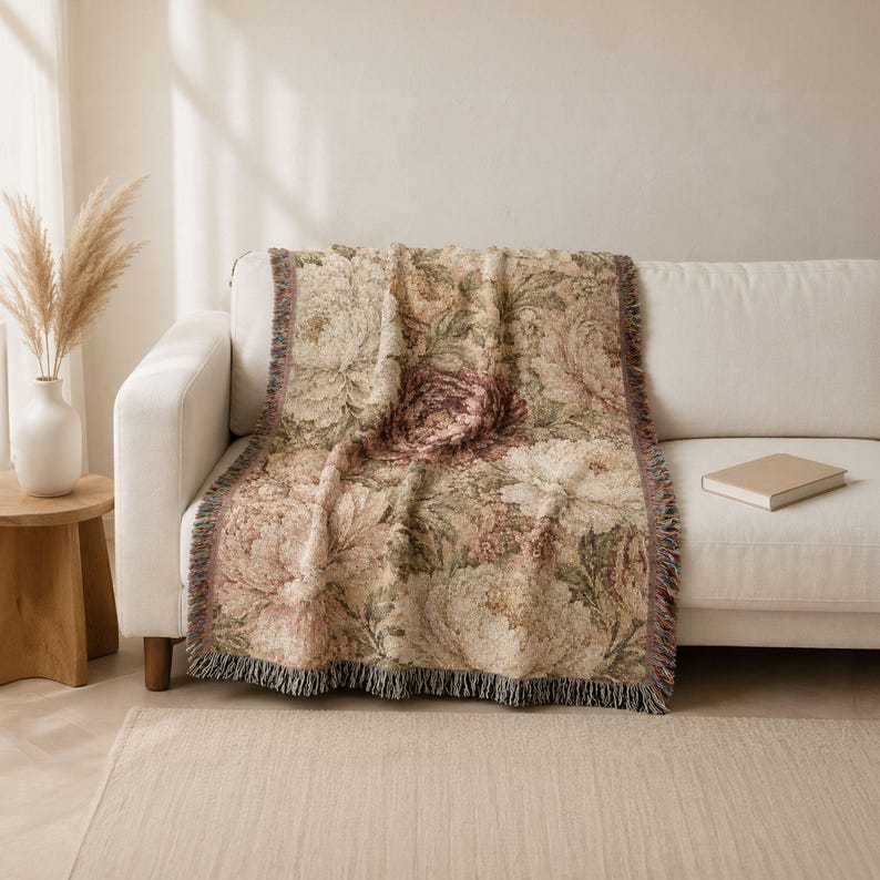 May include: A woven throw blanket featuring a detailed floral pattern with muted tones of cream, dusty pink, and sage green. The rectangular textile has a textured surface and finished edges with short, multi-colored fringe along the bottom border.
