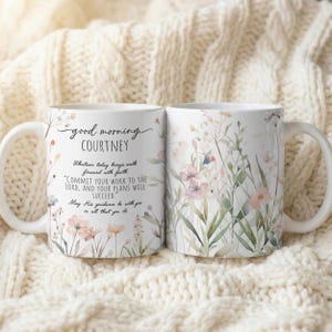 Op de afbeelding: Twee witte keramische mokken met bloemmotieven en de tekst "good morning COURTNEY". Eén mok heeft een citaat, terwijl de andere een bloemmotief heeft. De mokken staan tegen een crèmekleurige, gebreide achtergrond.