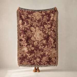Puede incluir: Una manta tejida rectangular con un fondo de color burdeos intenso y un patrón floral beige repetitivo de rosas y hojas. La manta tiene una apariencia texturizada y está terminada con un borde de flecos deshilachados en los cuatro lados.