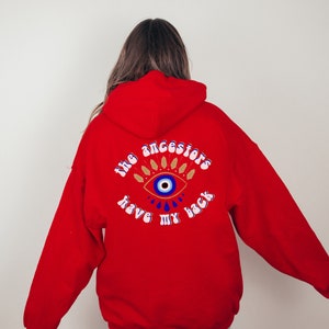 May include: Red hooded sweatshirt with a blue and white evil eye graphic and the text "the ancestors have my back" on the back.