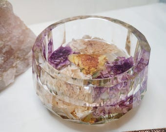 May include: A clear, faceted resin bowl filled with dried flowers in shades of purple, yellow, and cream. The bowl is round and sits on a white surface. A piece of rose gold trim with a geometric pattern is in the foreground.
