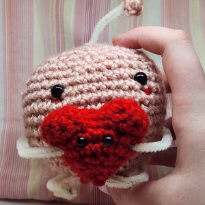 May include: A crocheted pink and red heart-shaped plush toy with black eyes. The toy has a white crocheted antenna and is holding a red heart.