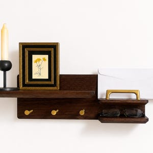 May include: A dark wood wall-mounted organizer featuring a shelf, three brass hooks, and a smaller side shelf with a brass handle. A black candle holder with a cream candle, a framed floral print, a white envelope, and eyeglasses are displayed.