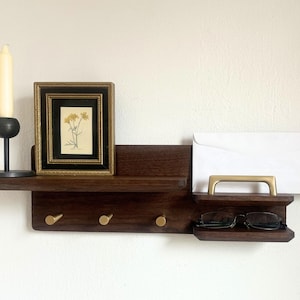 May include: A dark wood wall shelf with a candle, framed artwork, and a section for mail and glasses. The shelf has brass hooks and a brass handle. The artwork features a floral print.
