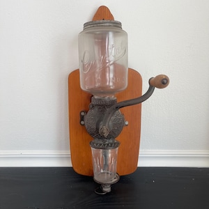 May include: An antique coffee grinder mounted on a wooden board. The grinder features a clear glass jar with the word "Crystal" and "No. 2" printed on it. The grinder has a black metal body with a wooden handle and a clear glass cup at the bottom.