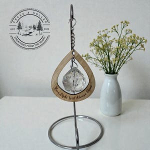 May include: A wooden teardrop-shaped ornament with a clear crystal ball suspended inside. The ornament hangs from a metal chain and stand. The text "Your Light Will Always Shine" is engraved on the wood. The logo "Creek & Holler" is in the upper left.