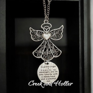 May include: A silver angel charm with a heart in the center and a round tag hanging below. The tag has the text "Guardian Angel Protect me, my passengers, and all who I pass by with a steady hand and a watchful eye" and the text "Creek and Holler".