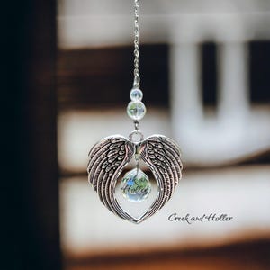 May include: A silver heart-shaped charm with angel wings and a clear crystal hanging from a silver chain. The crystal has the words "Creek and Holler" inscribed on it.