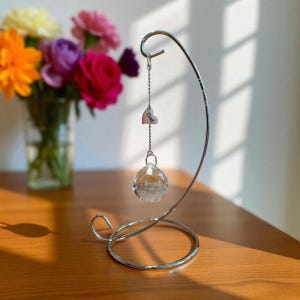 May include: A silver-colored metal hanging display stand with a heart-shaped charm that says "Dad" and a clear crystal ball. A vase of colorful flowers sits in the background on a wooden surface.
