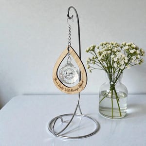 May include: A clear crystal ornament with a mountain scene inside, suspended from a wooden teardrop frame. The frame has the text "Your Light Will Always Shine" engraved on it. The ornament hangs from a silver metal stand.