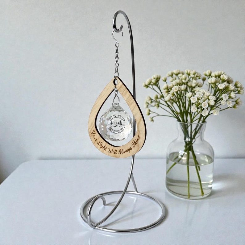 May include: A clear crystal ornament with a mountain scene inside, suspended from a wooden teardrop frame. The frame has the text "Your Light Will Always Shine" engraved on it. The ornament hangs from a silver metal stand.