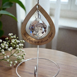 May include: A crystal ball ornament hangs within a teardrop-shaped wooden frame. The frame is engraved with the words "Your Light Will Always Shine." The ornament is suspended by a silver metal stand. The background includes white flowers and a window.
