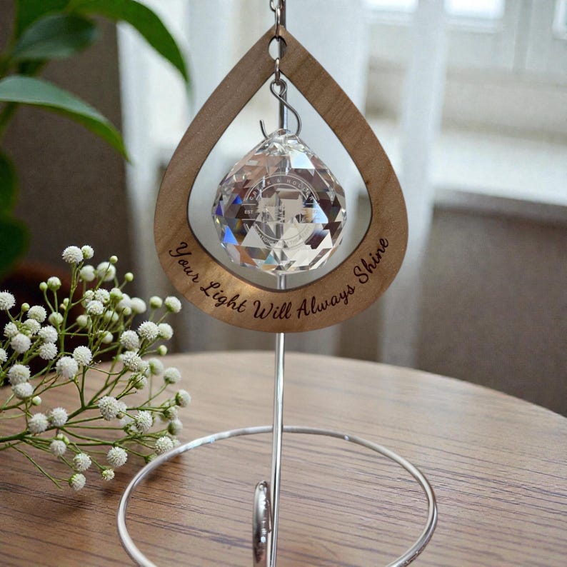 May include: A crystal ball ornament hangs within a teardrop-shaped wooden frame. The frame is engraved with the words "Your Light Will Always Shine." The ornament is suspended by a silver metal stand. The background includes white flowers and a window.