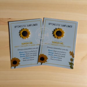 May include: Two light blue cards with a sunflower pin and text that reads "Optimistic Sunflower" and "Sending you sunshine, happy thoughts & good vibes." The cards have a wooden background.