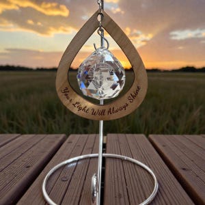 May include: A crystal ball ornament hangs within a wooden teardrop frame, inscribed with "Your Light Will Always Shine." The ornament is suspended by a silver metal stand. The background features a sunset over a field.
