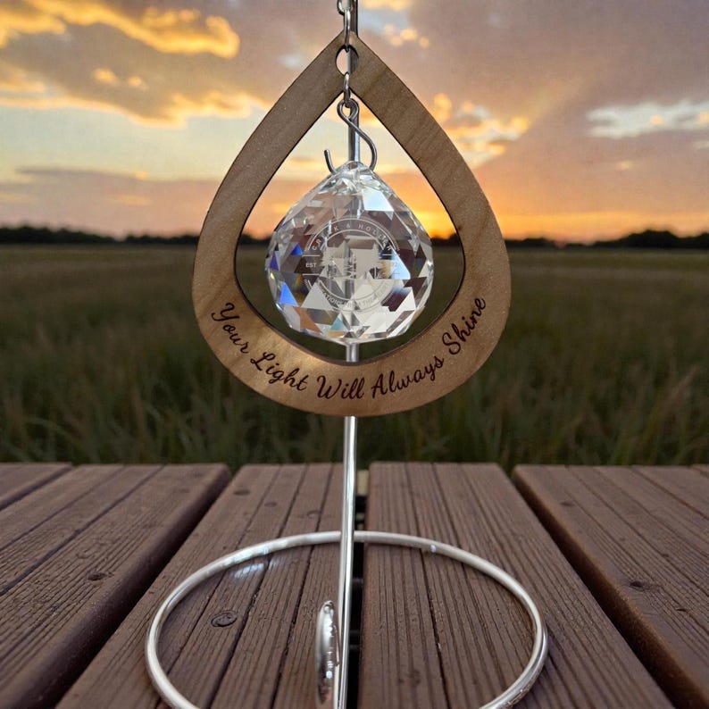 May include: A crystal ball ornament hangs within a wooden teardrop frame, inscribed with "Your Light Will Always Shine." The ornament is suspended by a silver metal stand. The background features a sunset over a field.