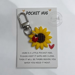 May include: A small sunflower keychain with a ladybug charm attached. The keychain is on a white card with the text "Pocket Hug" and a message about keeping it safe and close.
