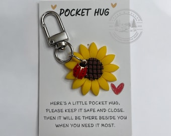 May include: A small sunflower keychain with a ladybug charm attached. The keychain is on a white card with the text "Pocket Hug" and a message about keeping it safe and close.
