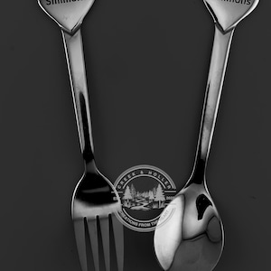 May include: A set of two silver heart-shaped spoons with the name "Simmons" engraved on each. The spoons are a fork and a spoon, and they are both polished to a shine.
