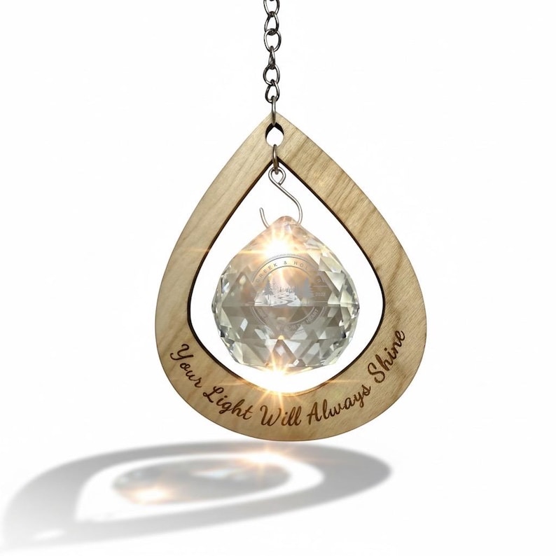 May include: A clear crystal prism hangs inside a teardrop-shaped wooden frame. The frame has the engraved text "Your Light Will Always Shine". A silver chain suspends the ornament, which casts a bright light.