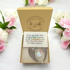 May include: A small, silver-colored angel wing ornament with a clear iridescent crystal ball hanging from it. The ornament is in a brown box with a white card that reads "There are some who bring a light so great to the world that even after they have gone, the light remains."