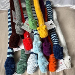 Può includere: Una collezione di lunghi serpenti all'uncinetto in peluche in vari colori solidi e motivi a righe, tra cui rosso, giallo, verde, blu, viola, arancione, grigio e bianco. Ogni serpente presenta piccoli occhi neri e un'etichetta bianca.