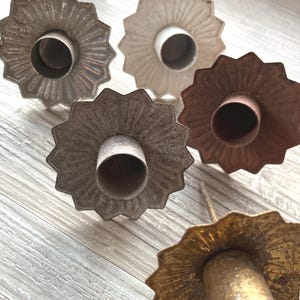 Könnte beinhalten: Eine Sammlung von Vintage-Kerzenhaltern in verschiedenen metallischen Ausführungen. Die Halter haben ein Sternmuster um die Basis und ein zentrales Rohr für die Kerze. Farben: Silber, Off-White, Braun und Gold.