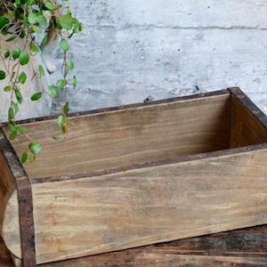 Könnte beinhalten: Eine rechteckige Pflanzkasten aus Holz mit Metallakzenten. Die Box hat eine natürliche Holzoberfläche und ist für Pflanzen konzipiert. Der Pflanzkasten steht auf einer Holzoberfläche mit einem Steintopf und Grünzeug im Hintergrund.