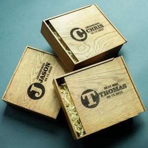 Könnte beinhalten: Drei Holzkisten mit Schiebedeckeln, jeweils mit Namen, Initialen und Datum graviert. Die Kisten sind mit Stroh gefüllt und aus hellem Holz gefertigt. Der Text auf den Kisten lautet "Groomsman Chris 09.15.2025", "Groomsman Jason 09.15.2025" und "Best Man Thomas 09.15.2025".