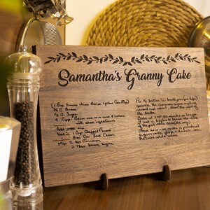 K&ouml;nnte beinhalten: Ein Schneidebrett aus Holz mit einem handgeschriebenen Rezept f&uuml;r Samanthas Oma-Kuchen. Das Rezept enth&auml;lt Zutaten wie Butter, Zucker, Eier und Pekann&uuml;sse. Das Brett hat einen dekorativen Rand aus Bl&auml;ttern.