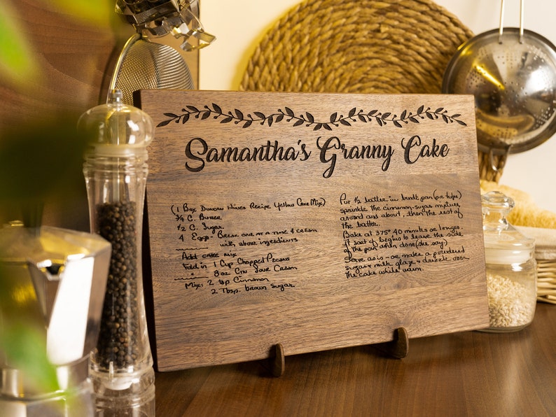 K&ouml;nnte beinhalten: Ein Schneidebrett aus Holz mit einem handgeschriebenen Rezept f&uuml;r Samanthas Oma-Kuchen. Das Rezept enth&auml;lt Zutaten wie Butter, Zucker, Eier und Pekann&uuml;sse. Das Brett hat einen dekorativen Rand aus Bl&auml;ttern.