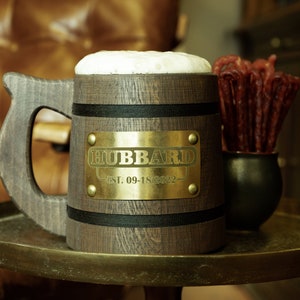 May include: A wooden beer stein with a black band and a gold plaque that reads "HUBBARD EST. 09-18-2022". The stein is filled with a foamy beverage and is sitting on a gold-colored table.