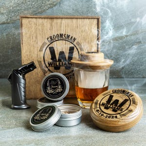 May include: A wooden box with the word "Groomsman" and the letter "W" engraved on it. The box contains a set of whiskey stones, a glass, a wooden lid, and a torch. The box is on a gray surface.