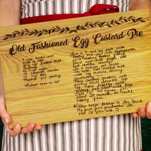K&ouml;nnte beinhalten: Ein Schneidebrett aus Holz mit einem handgeschriebenen Rezept f&uuml;r einen altmodischen Eier-Custard-Kuchen. Das Rezept enth&auml;lt Zutaten und Anweisungen zum Backen des Kuchens. Das Brett ist hellbraun mit einer nat&uuml;rlichen Holzmaserung.