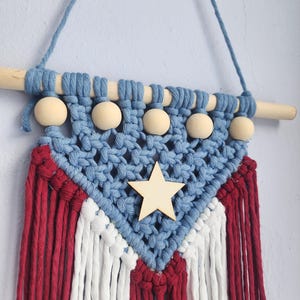 May include: A macrame wall hanging with a patriotic theme. It features a blue top section with wooden beads and a wooden star. The lower section has red and white fringe, creating a flag-like design.
