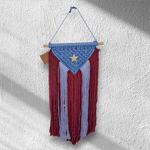 May include: A blue and red macrame wall hanging with a white star in the center. The hanging is made of yarn and has a wooden dowel at the top.